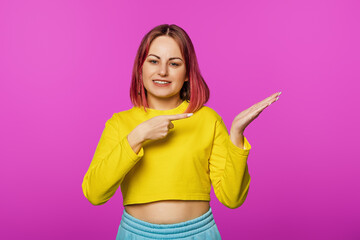 Obraz na płótnie Canvas Happy smiling young woman with pink hair looking at camera and pointing with finger at copy space on pink background studio
