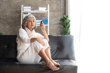Depressed Asian senior woman sit on sofa looking away by the window. Loneliness elderly female living alone at home with sadness face. Retirement mental health care and illness recovery concept