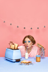 Beautiful young girl with bright makeup drinking milk, eating toast with confetti over pink studio...