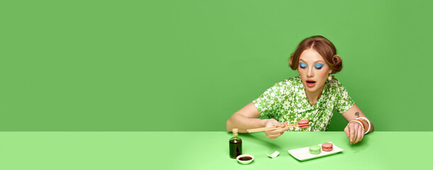 Stylish young girl eating macarons with soy sauce over green studio background. Weird taste. Food pop art photography. Concept of retro style, creative vision, imagination. Banner. Copy space for ad