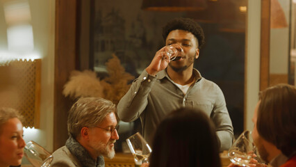 African American man says toast. Group of friends clink glasses and drink wine in restaurant. People spend evening together, hangout on birthday party in modern gastro cafe. Public catering concept.