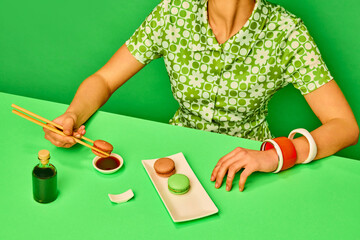 Cropped image of girl putting sweet macarons into soy sauce with chopsticks against green background. Extraordinary taste combination. Food pop art photography. Concept of retro style, creative vision