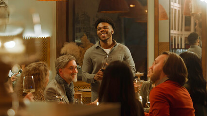 African American man says toast. Group of friends clink glasses and drink wine in modern gastro cafe. People spend evening together, hangout on birthday party in restaurant. Public catering concept.