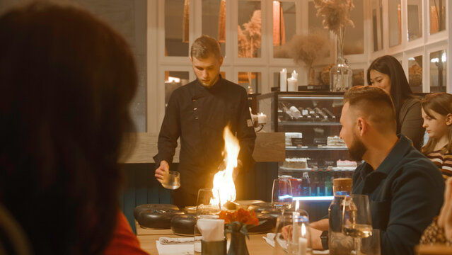Male chef prepares meal in flambe style in front of people in modern gastro cafe. They film the process on their phones. Group of friends spend weekend evening in restaurant. Concept of public eating.