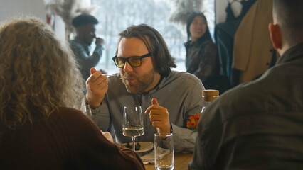 Caucasian man sits in modern gastro cafe, eats delicious dish, drinks wine and talks to friends. Group of friends celebrate birthday or spend weekend together in restaurant. Concept of public eating.