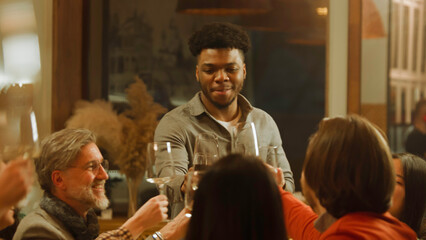 African American man says toast. Group of friends clink glasses and drink wine in restaurant. People spend evening together, hangout on birthday party in modern gastro cafe. Public catering concept.