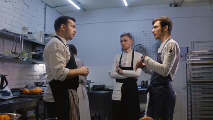 Adult chef stands with colleagues and discussing menu changes. Coworker prepares dish with vegetables at background. Modern kitchen illuminated by lamps. Concept of working at restaurant. Slow motion.