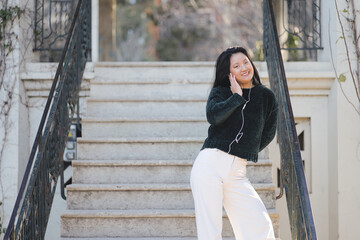 Attractive young Asian girl happily listening to music with headphones from music app on mobile phone, on steps of a park.