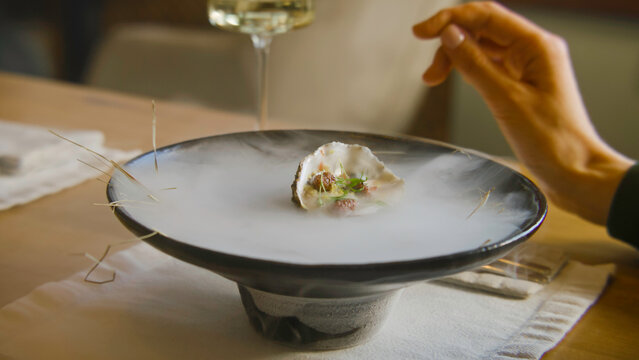 Close up shot of pouring liquid in meal to make dry ice steam for dish presentation in molecular cuisine restaurant. Hand of woman sitting at the table in modern gastro cafe. Concept of public eating.