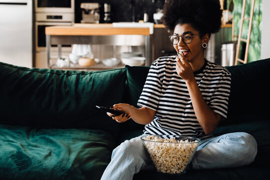 Cheerful afro woman watching tv and eating popcorn while sitting on couch