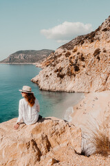 Woman wearing a hat sitting on the rock watching beautiful sandy beach below.