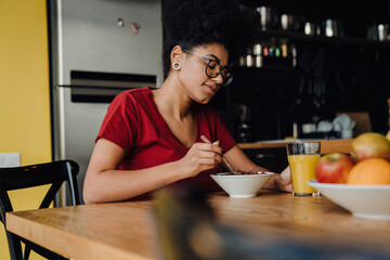 Beautiful african woman drinking orange juice and eating cereal while sitting in kitchen