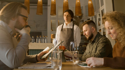 Male waiter comes to the group of friends or relatives and talks with people. Guests watching menu and making order. Celebrating anniversary, birthday dinner in gastro cafe. Concept of public eating.