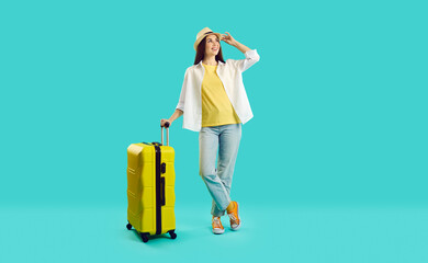 Full body happy young woman in summer hat, casual shirt and blue jeans standing with yellow suitcase isolated on turquoise background, looking away and smiling. Holiday, vacation, traveling concept - obrazy, fototapety, plakaty