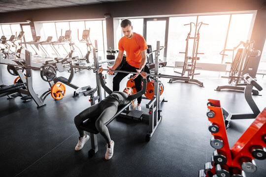 Young sporty girl works with barbell performing bench press. Personal trainer helps to perform the exercise. Wide angle shooting. - Powered by Adobe