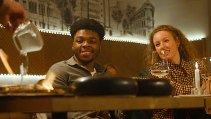 The chef prepares meal in flambe style in front of people in modern gastro cafe. Cheerful multi ethnic friends spend weekend evening, hangout on birthday party in restaurant. Concept of public eating.