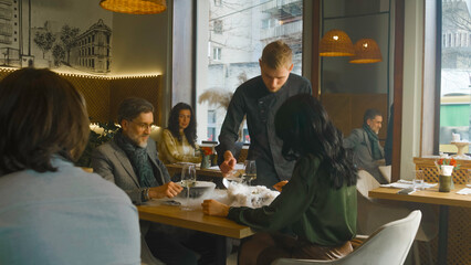 Adult man and woman sit at table in modern cafe together, drink wine and talk. Male waiter and chef...