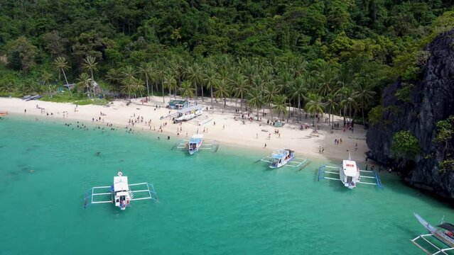 Aerial view: catamaran style Filipino banca boats traversing in foreground of Tropical Seven Commando beach , Tourists swimming in turquoise clear water on white sand .El Nido Palawan, Philippines.