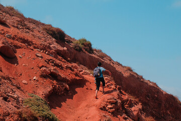 Fototapeta na wymiar Sidi Ifni, Morocco - local boy with backpack walks barefoot up a hill. Young Moroccan hikes to school. View from behind. Blue day sky. Red clay soil. Youth travel, explore, adventure concept.