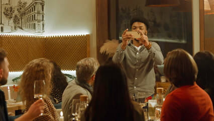 African American man takes photo and selfie with friends in cozy cafe using phone. Group of diverse people spend evening together, hangout on birthday party in restaurant. Concept of public eating.
