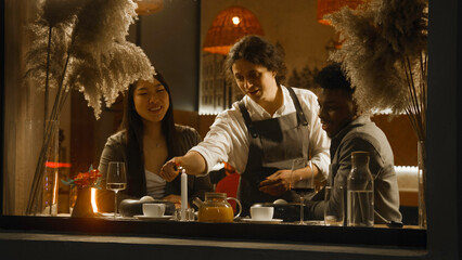 African american man and asian woman sit in modern cafe together, drink wine and talk. Male waiter brings dishes, lights candle. Multi ethnic couple on a date in restaurant. Concept of public eating.