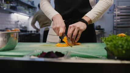 Hands close-up of professional male cook standing by kitchen table and cutting sweet potato. Busy coworker prepares dishes at background. Concept of teamworking in restaurant. Slow motion. Dolly shot.