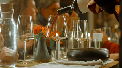 Close up shot of waiter pouring red wine from the bottle into the glass in restaurant. People clink glasses, celebrate birthday, spend weekend evening in modern gastro cafe. Public catering concept.