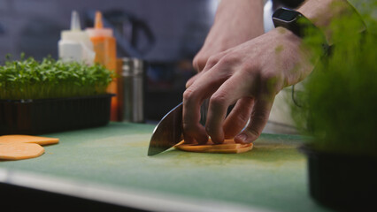 The cook cuts sweet potato on cutting board. Hands of professional chef close-up. Greenery and other ingredients on kitchen desk. Process of cooking gourmet dish. Public catering. Vegetarian dish.