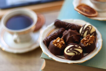 Books, reading glasses, e-reader, plate of chocolate pralines, bowl of cookies, cups of tea and lit candles on the table. Selective focus.