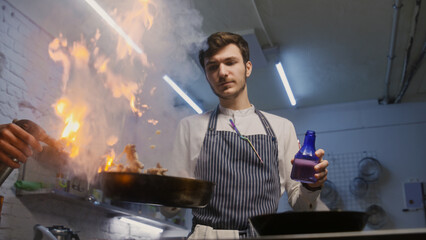 Male chef in apron sets fire to frying pan, fries beef meat in flambe style using gas burner and adds pepper. Process of preparing gourmet dish. Restaurant with professional cuisine. Gastronomic art.