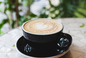 Black cup of cappuccino with latte art of heart shape on saucer on concrete background with tropical plant. Breakfast drink