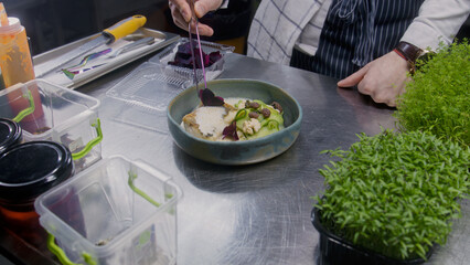 The chef makes presentation of dish. Male cook puts decorative flower petals on top of fried fish and steamed vegetables in white bechamel sauce. Restaurant with gourmet cuisine. Public catering.