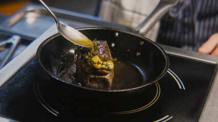 Chef cooks tasty meal on stove. Male cook fries juicy meat steak on pan with rosemary and garlic, pours beef meat with oil. Process of roasted steak cooking. Restaurant with professional cuisine.