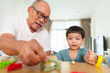 Portrait of smiling Asian Senior man and Little child boy molding modeling clay together. Grandfather and Grandson making toys from plasticine. Creativity and hobby.