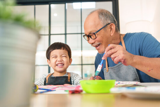 Cheerful Asian Japanese Senior man and little child boy in apron Enjoying painting art at home together. Happy Grandfather and Grandchild playing and laughing. Positive emotion.