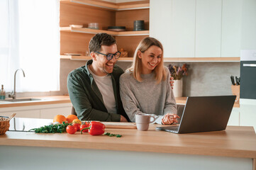 Sitting, using laptop. Young couple is on the kitchen together
