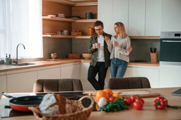 Standing, talking, holding cups with drink. Young couple is on the kitchen together