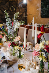 Festive table decorated with composition of red, pink flowers and greenery, candles in hall. Table newlyweds and guest in banquet area on wedding party. Luxurious celebration of event in restaurant.