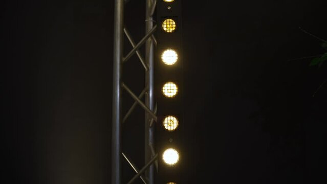 Stage lighting fixture is attached to vertical metal truss, white spotlight illuminating dark wall from behind. Halogen bulbs behind bars alternately dim and glow warm.