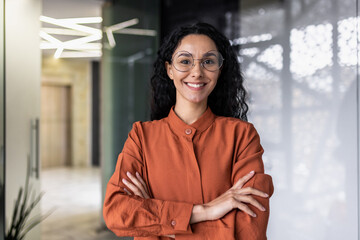 Portrait of happy and successful business woman, boss in shirt smiling and looking at camera inside office with crossed arms, Hispanic woman with curly hair in corridor - obrazy, fototapety, plakaty