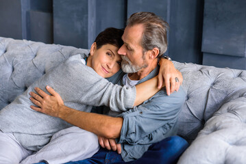 An elderly couple sitting on a gray sofa in an embrace. Home comfort and coziness.