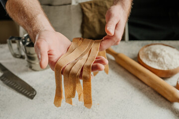 Unrecognizable man in an apron holding dough cut into strips on a white table against a dark wall © lithiumphoto