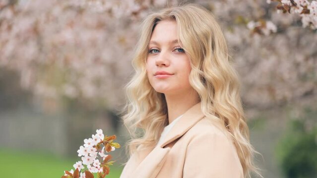 Close-up portrait of a stunning woman with blond hair looking at the camera smiling outdoors against the background of blooming trees in spring. Beautiful blooming cherry tree on the background.