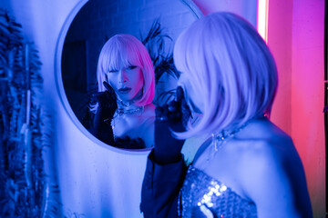 Fashionable drag queen in wig looking at mirror in neon light at home.