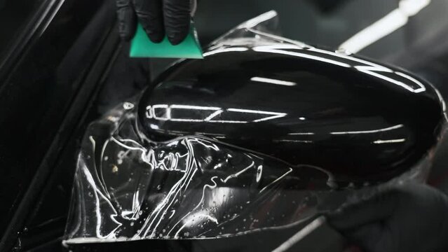 Close-up of a hand in a balck glove using a green scraper ti smoothen paint protection film put onto a car mirror. High quality 4k footage