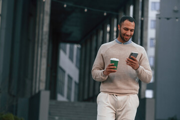 With cup of coffee and smartphone. Handsome black man is outdoors near the business building