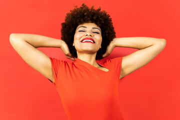 Obraz na płótnie Canvas A dark-skinned woman with afro hair posing on a red background. The girl has a relaxed expression with her eyes closed and her hands behind her head. Concept of relaxation, dreaming, rest or positive.