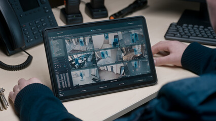 Security officer controls CCTV cameras in coworking office, uses digital tablet with surveillance cameras playback on screen. High tech security with facial recognition. Monitoring and social safety.