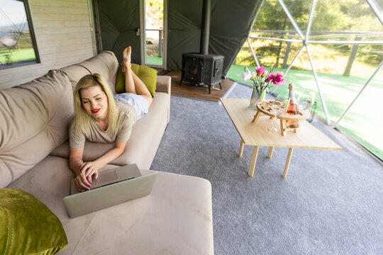 Woman working on laptop geo dome tents. Green, blue background. Cozy, camping, glamping, holiday, vacation lifestyle concept. Outdoors cabin, scenic background.