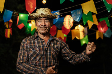 Senior man celebrating the Brazilian Festa Junina. Portrait of a man wearing typical clothes and a straw hat under the decoration of pennants and lights from the traditional June festival in Brazil. - obrazy, fototapety, plakaty